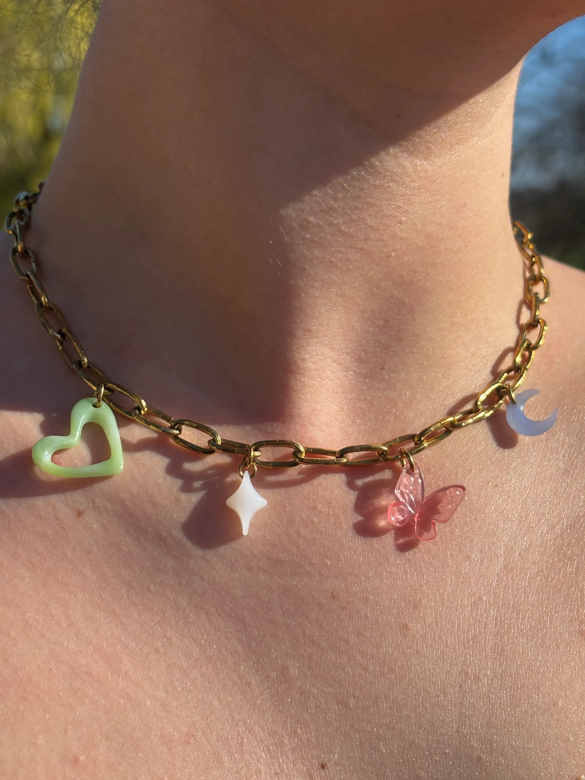 Collier doré gros maillon quatre charms. Un grand cœur vert organique, une étoile blanche, un papillon rose, une lune bleue